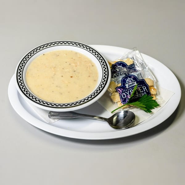 A bowl of creamy soup with a checkered rim, oyster crackers, and a spoon on a white plate.